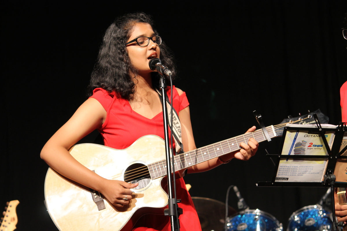 Student singing into a microphone while playing acoustic guitar at a music recital